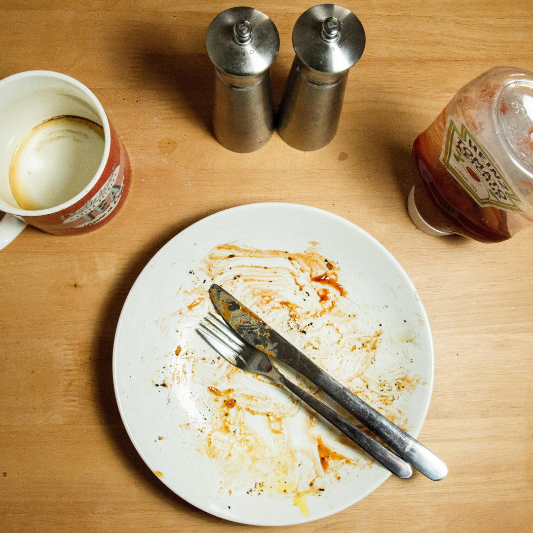 En vit tom mattallrik med en gaffel och en kniv som ligger för att signalera att maten är uppäten. En ketchupflaska, salt- och pepparkar och en tom mugg står runt tallriken. 